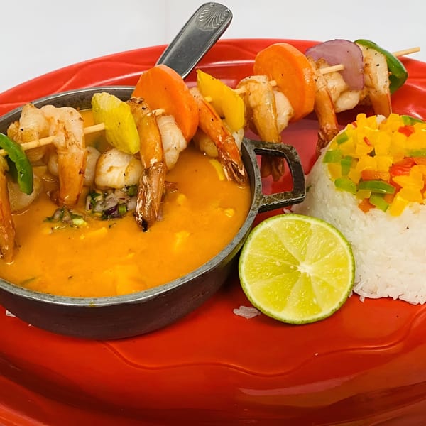 Peruvian shrimp skewers with vegetables, orange sauce, white rice, and a lime wedge on a red plate.