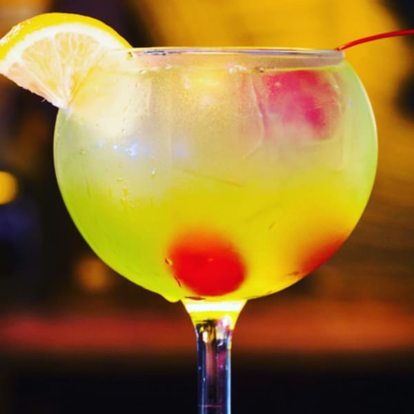 A vibrant yellow cocktail with a lemon slice and two red cherries in a large glass.