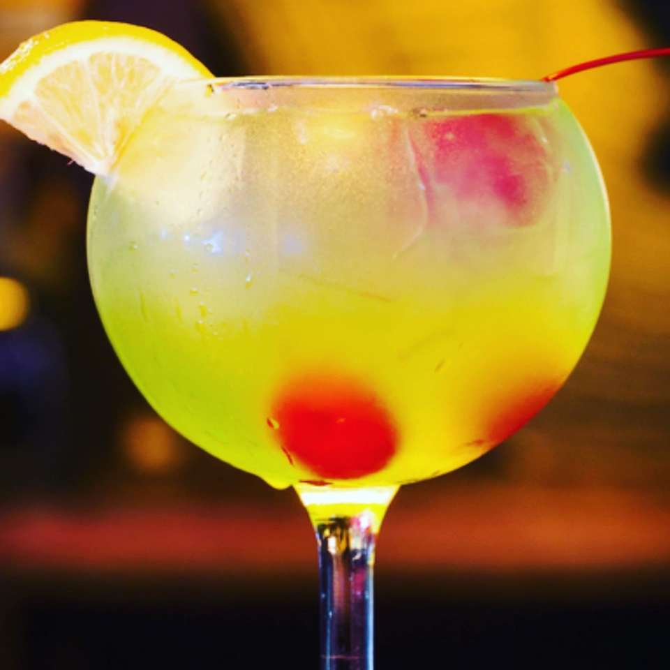 A vibrant yellow cocktail with a lemon slice and two red cherries in a large glass.