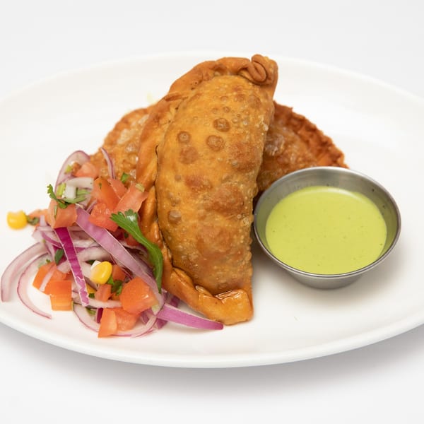 Two golden-brown Peruvian empanadas with a side of red onion salad and green dipping sauce.