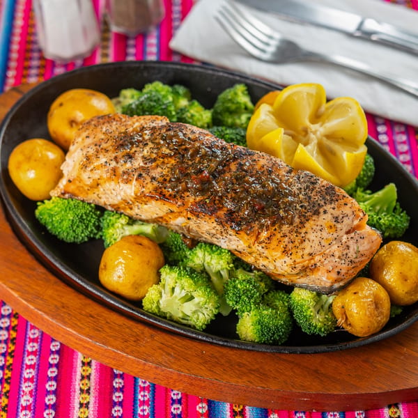Peruvian grilled salmon with roasted potatoes, broccoli, and lemon wedges on a sizzling platter.