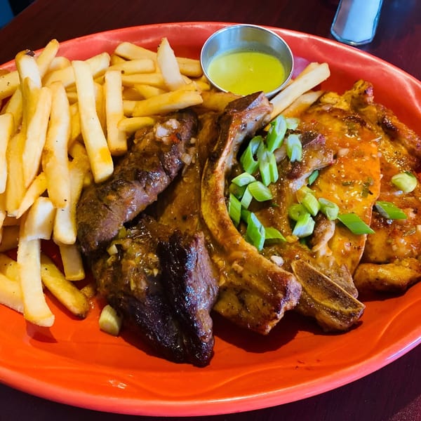 Peruvian grilled meats, including ribs and chicken, with french fries and a side of green sauce.