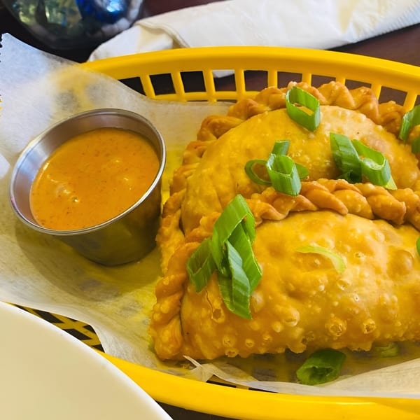 Golden Peruvian empanadas topped with green onions, served in a yellow basket with a side of orange sauce.